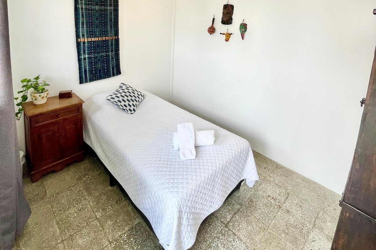 Una habitación pequeña y limpia con una cama individual, una mesita de noche, un estante en la pared y una ventana con cortina azul.