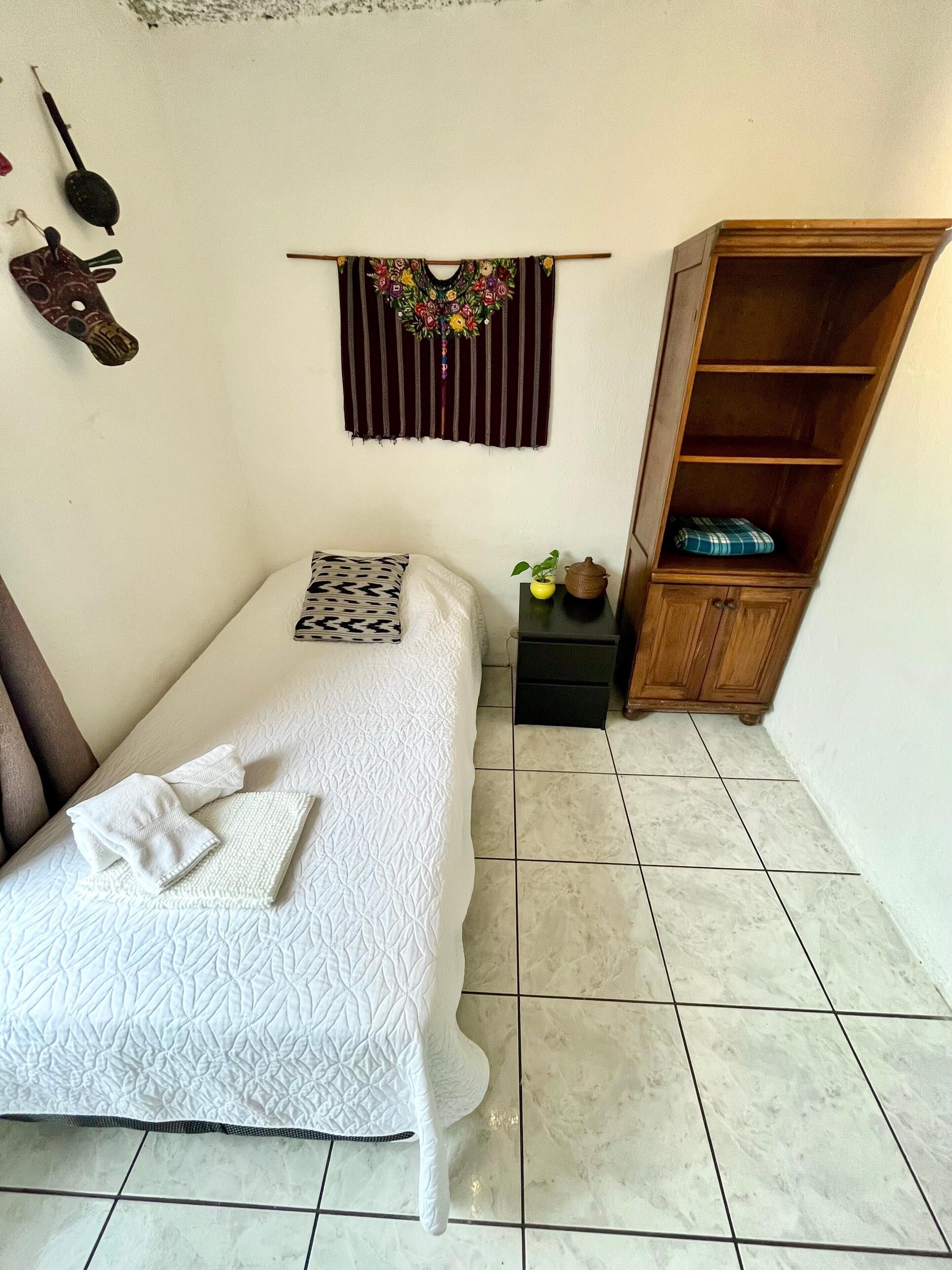 Une petite chambre à coucher, bien rangée, avec un lit simple, une table de chevet, une armoire en bois et un élément décoratif fixé au mur.