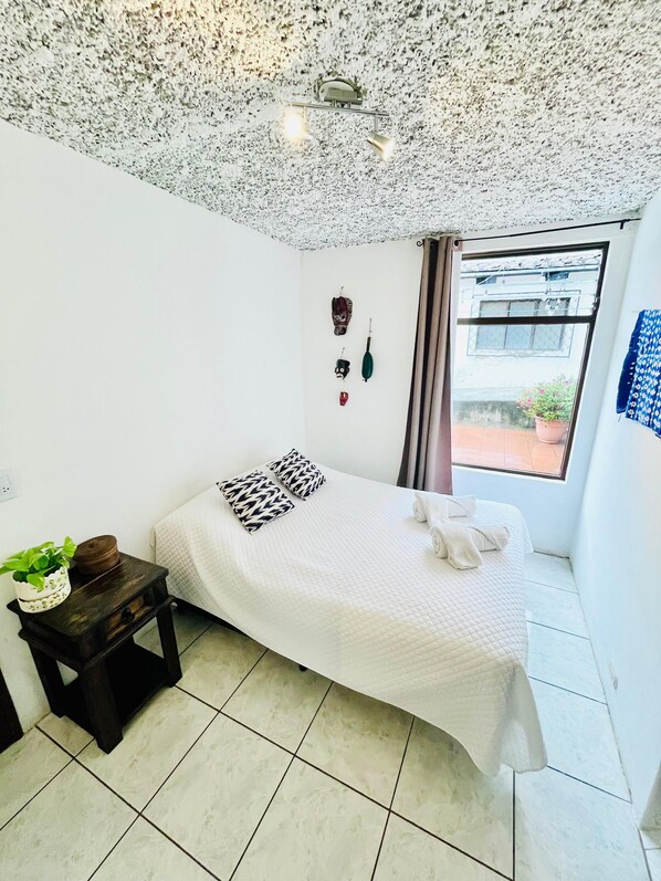 A bedroom with a bed, a small table with a plant, a window with curtains, and a ceiling with a light fixture. - Casa ELA Candelaria (Antigua Guatemala)