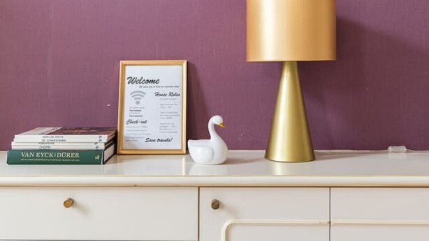 Un escritorio con un cartel de bienvenida enmarcado, una lámpara, una figurita de cisne blanco y una pila de libros frente a una pared morada.