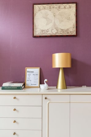 Ein Schreibtisch mit einem gerahmten Willkommensschild, einer Lampe, einer weißen Schwanfigur und einem Stapel Bücher vor einer violetten Wand.