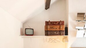 Una stanza piccola con pavimento in legno, un letto singolo, un comodino con una lampada, un televisore su uno scaffale e una sedia.