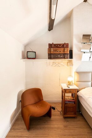 Una habitación pequeña con piso de madera, una cama individual, una mesita de noche con lámpara, un televisor en un estante y una silla.