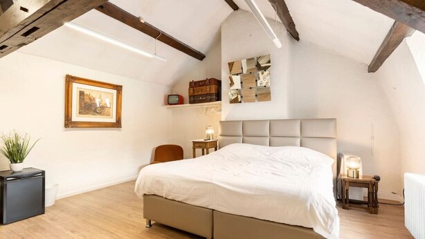 Una camera da letto con un grande letto, un comodino, una sedia, un tavolino con una lampada, un quadro appeso al muro e una mensola con oggetti decorativi.
