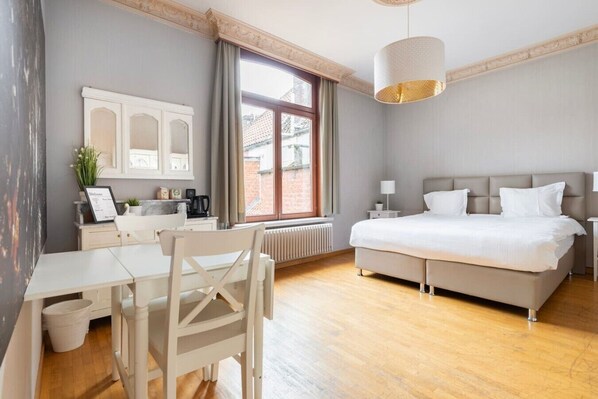 A hotel room with a bed, a desk, a chair, a radiator, and a window with curtains.