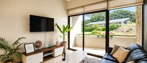 A modern living room with a flat-screen TV, a coffee table, a sofa, and a balcony with a view of greenery and a building.