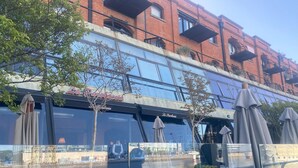 An outdoor café with tables and chairs, a glass wall reflecting the building behind, and a small tree in the foreground. - Puerto Madero Piso 20 Vista al Rio  (Buenos Aires)