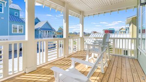 A covered porch with white railings and wooden flooring, overlooking a row of colorful houses. - Multi-level home with beach access, outdoor pool, outdoor shower, balcony (Port Aransas)
