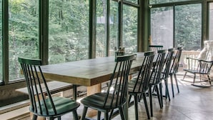 Un coin repas avec une table et des chaises en bois, de grandes fenêtres donnant sur la forêt et un fauteuil à bascule près de la fenêtre.