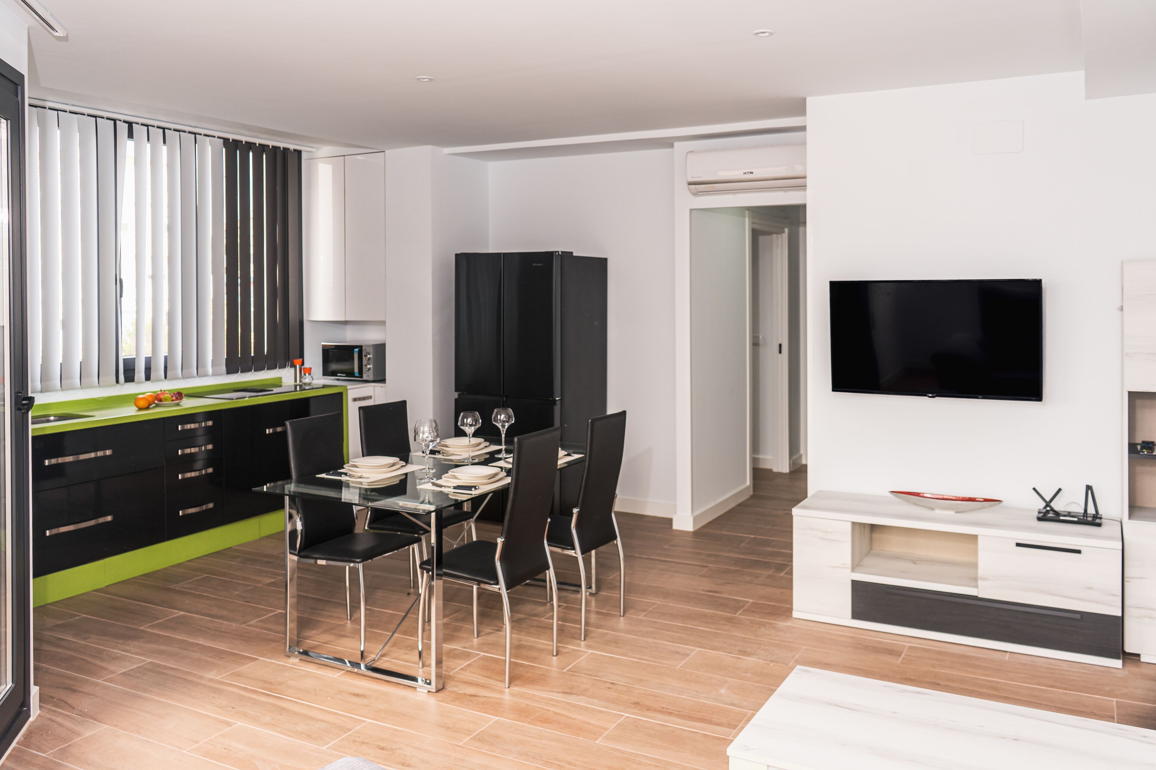 A modern kitchen with black cabinets, a dining table with chairs, and a flat-screen TV mounted on the wall.
