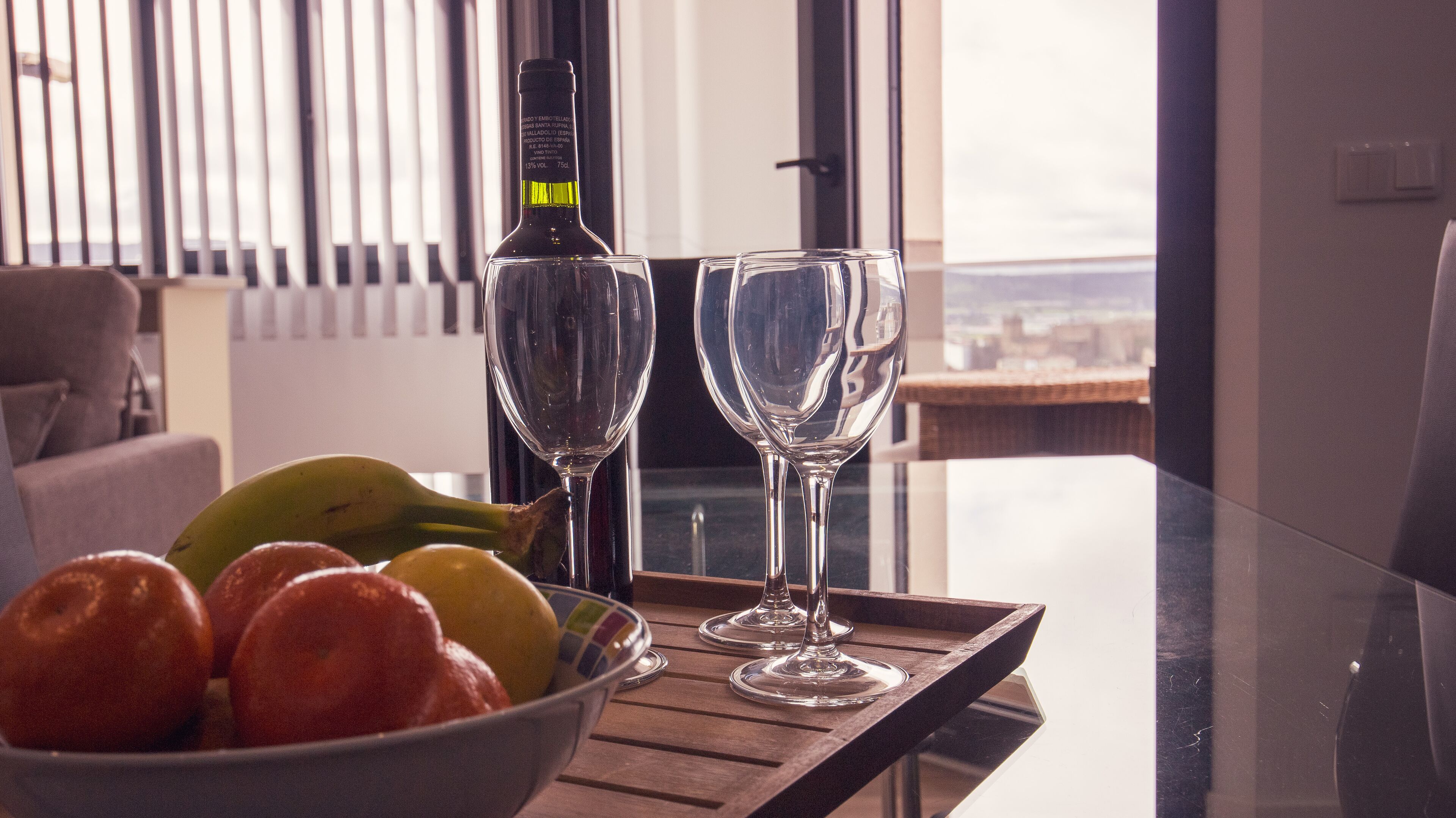 A table with a bottle, two glasses, and a bowl of fruit.