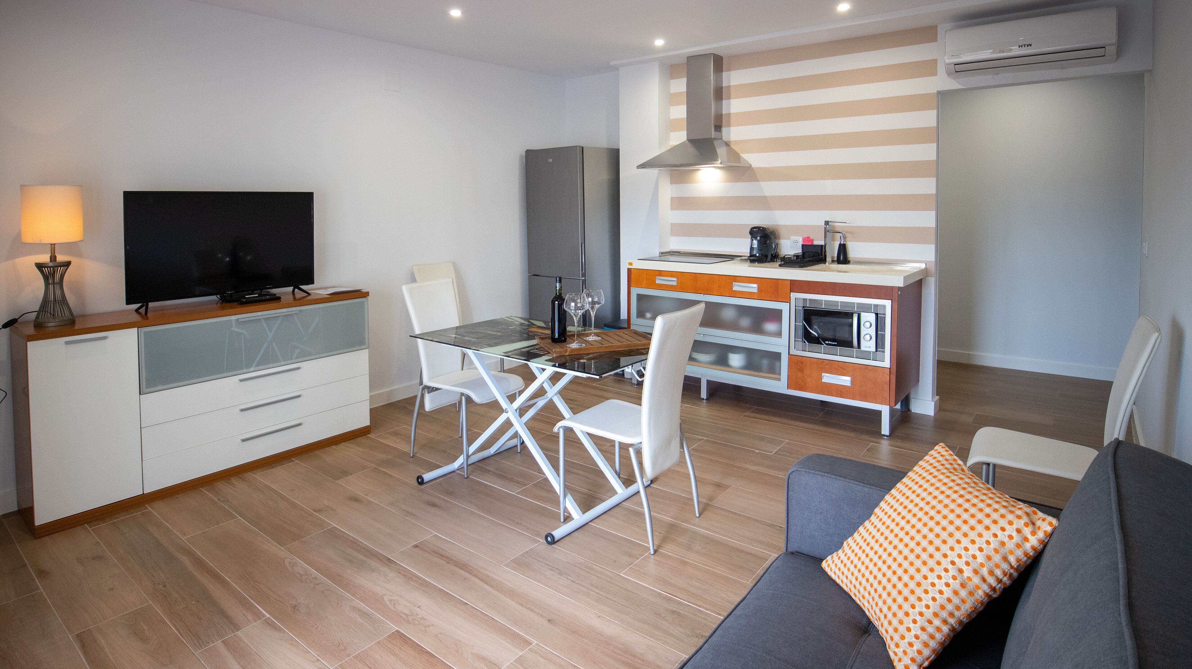 Una sala moderna con un sofá, televisor, mesa de comedor con sillas, una zona de cocina con microondas y una pared a rayas.