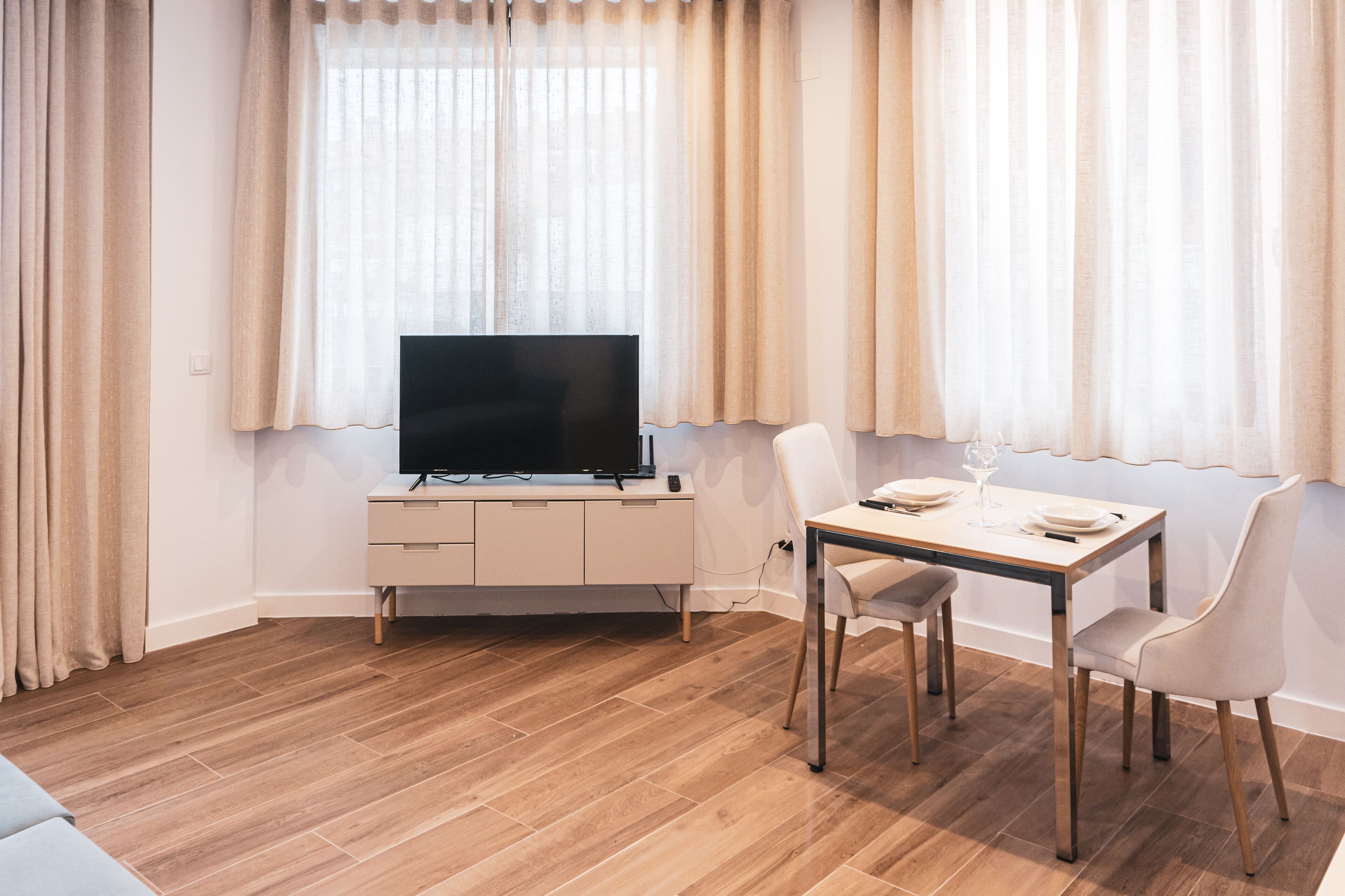 Een moderne woonkamer met een flatscreen televisie op een wit kastje, een eettafel met stoelen en een bank.