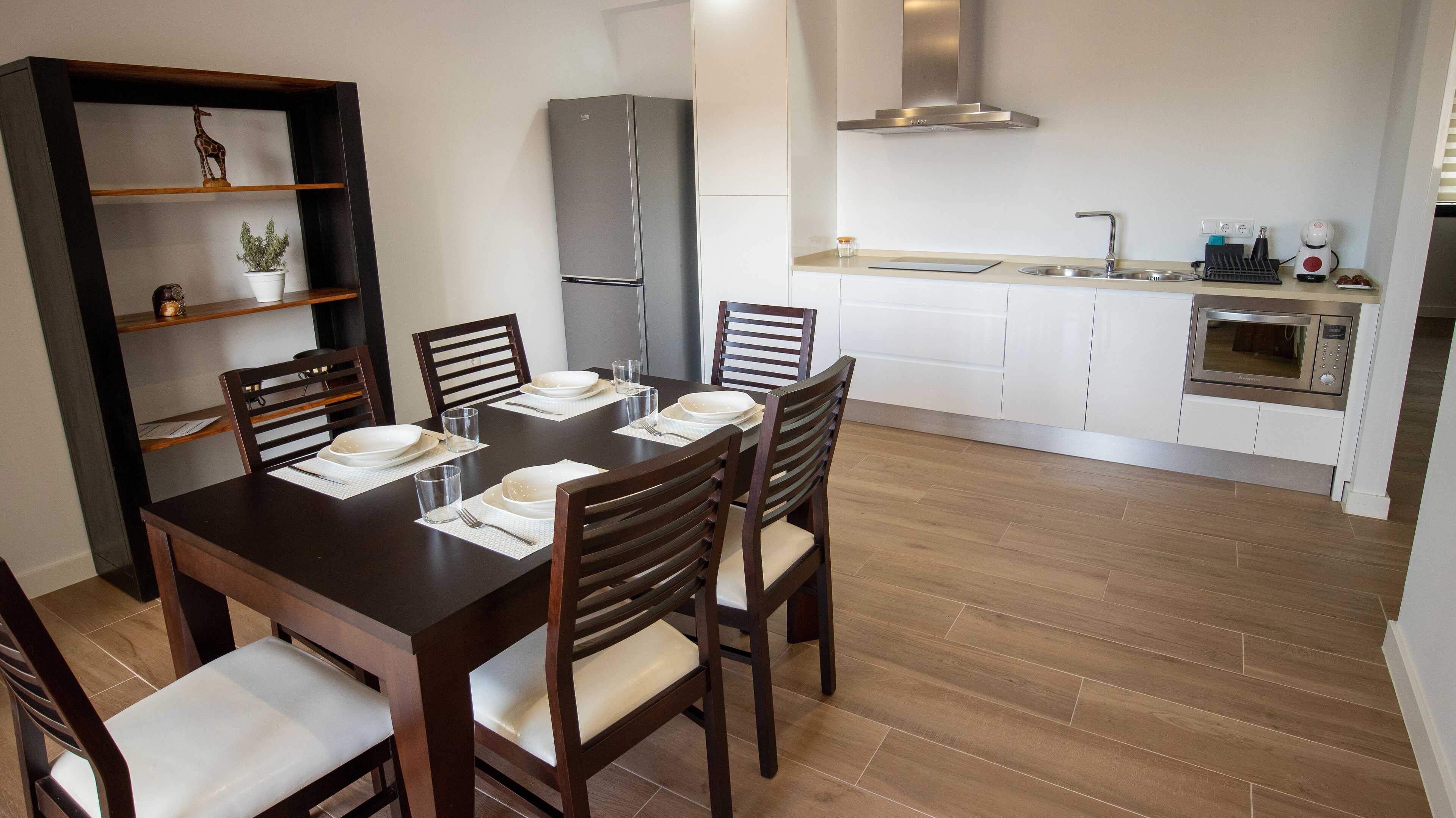 Un comedor con una mesa de madera dispuesta para cuatro personas, rodeada de sillas. Se ve una cocina en el fondo con armarios blancos y un horno empotrado.