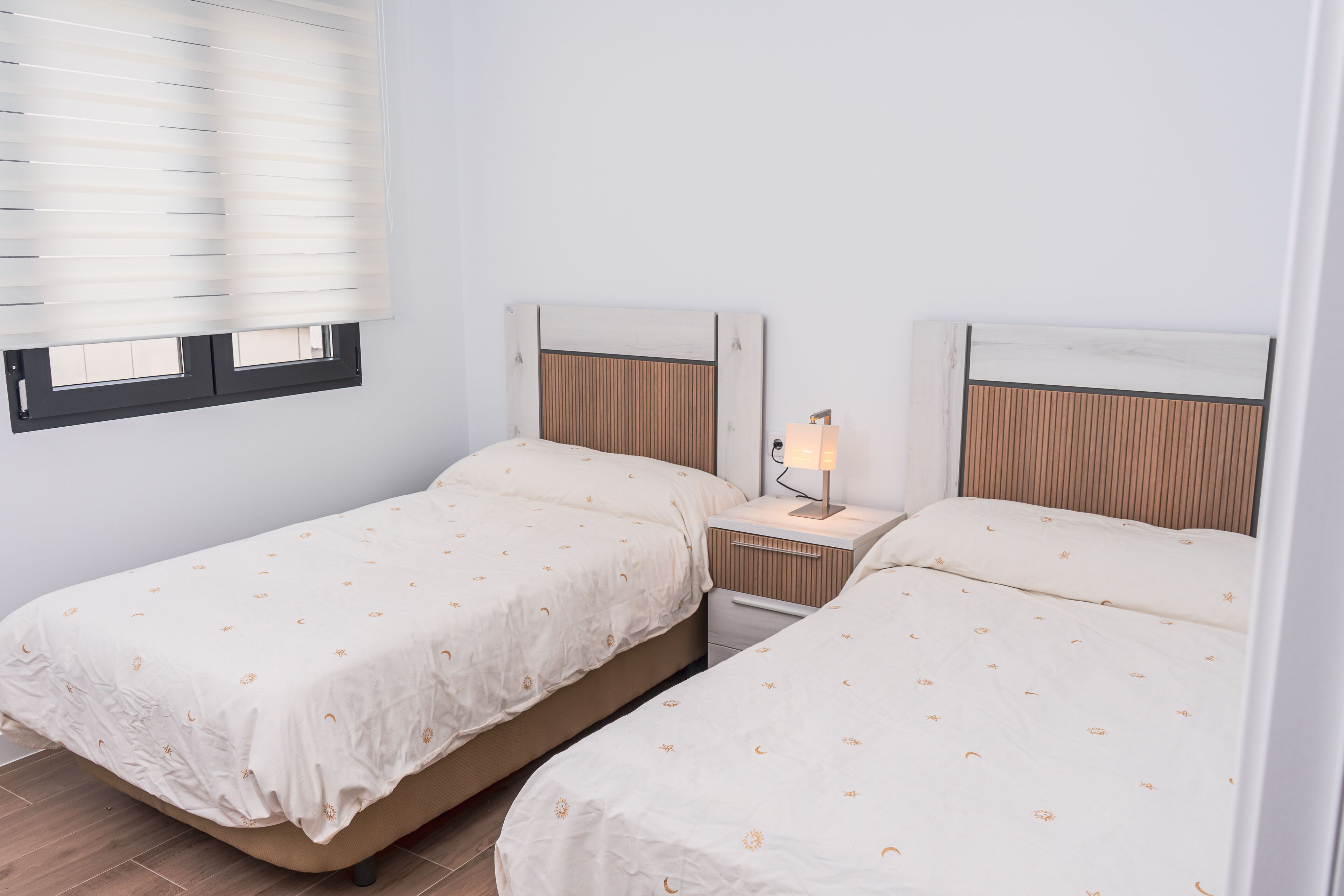 A twin room with two beds, a nightstand with a lamp, and a window with blinds.