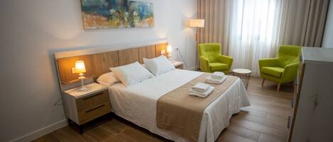 A hotel room with a large bed, two bedside tables with lamps, a painting on the wall, and two green armchairs.