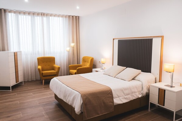 Habitación de hotel con cama, dos sillas, mesita de noche con lámpara, armario y ventana grande con cortinas.