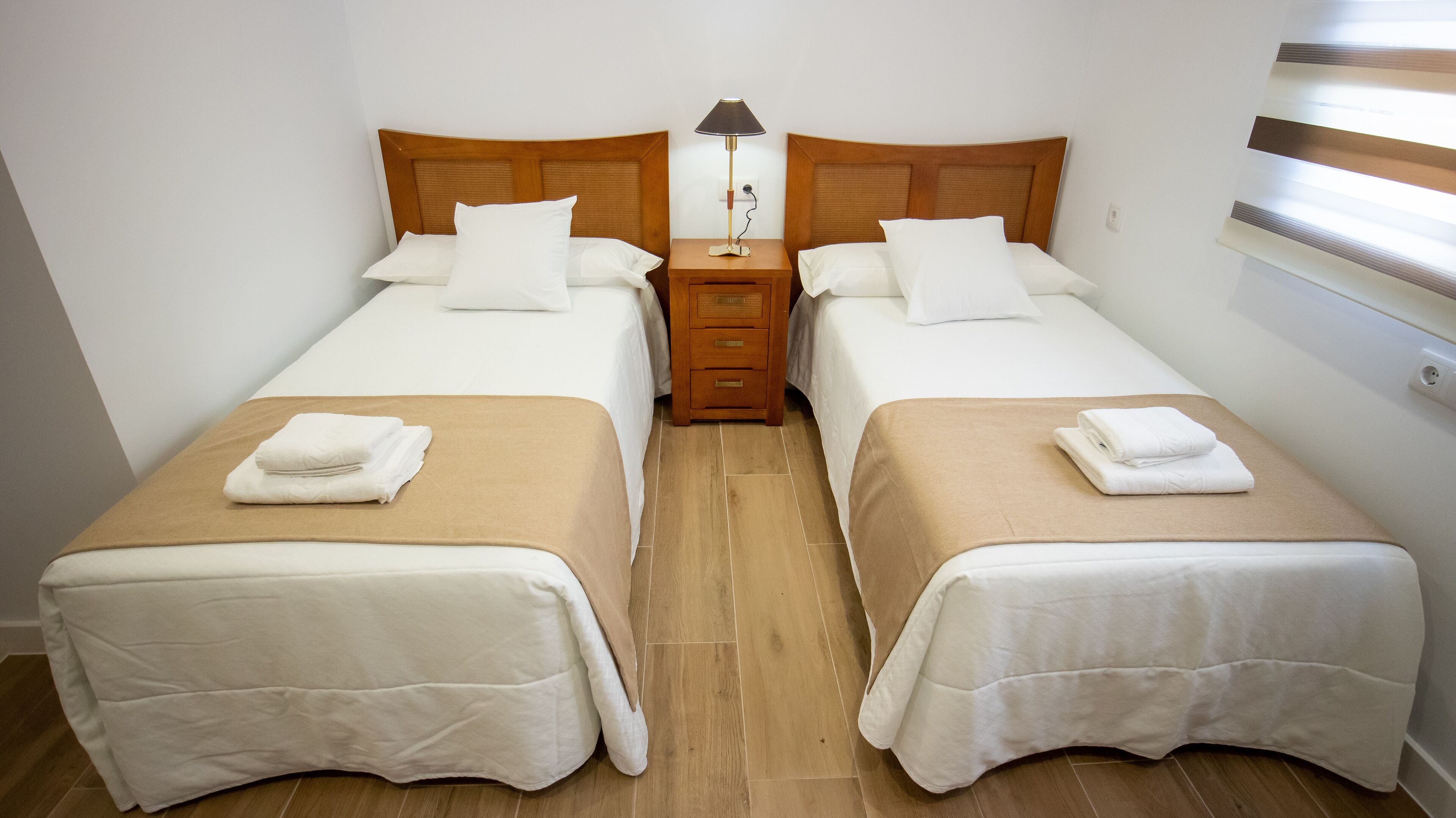 Ein Doppelzimmer mit zwei Betten, jeweils mit einem Holz-Kopfteil, einem Nachttisch dazwischen und einer Lampe auf dem Nachttisch.