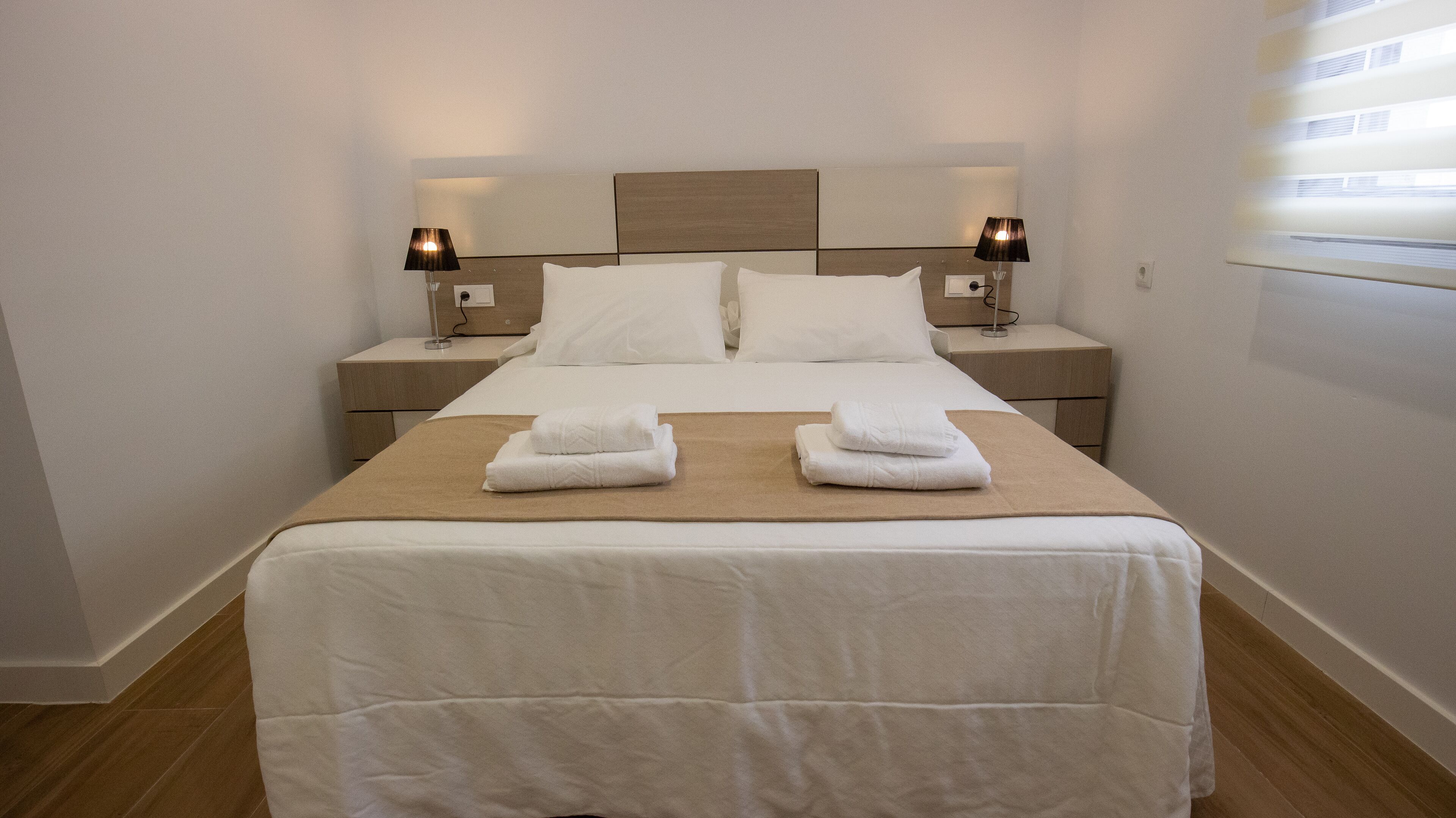 Una cama bien hecha con sábanas blancas y dos toallas dobladas sobre ella. Hay dos mesitas de noche con lámparas y un cabecero con un panel de luz.