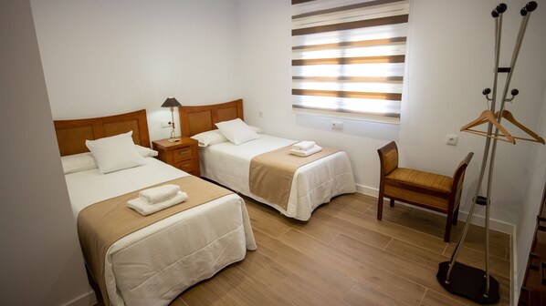Una habitación de hotel con dos camas, una mesita de noche de madera con lámpara, una silla de madera y un perchero.