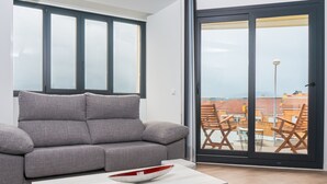 A modern living room with a grey sofa, a white coffee table, and a large sliding glass door leading to an outdoor area with a view of buildings and a street lamp. - Alojamientos VEN, Vista Extremadura Norte (Plasencia)