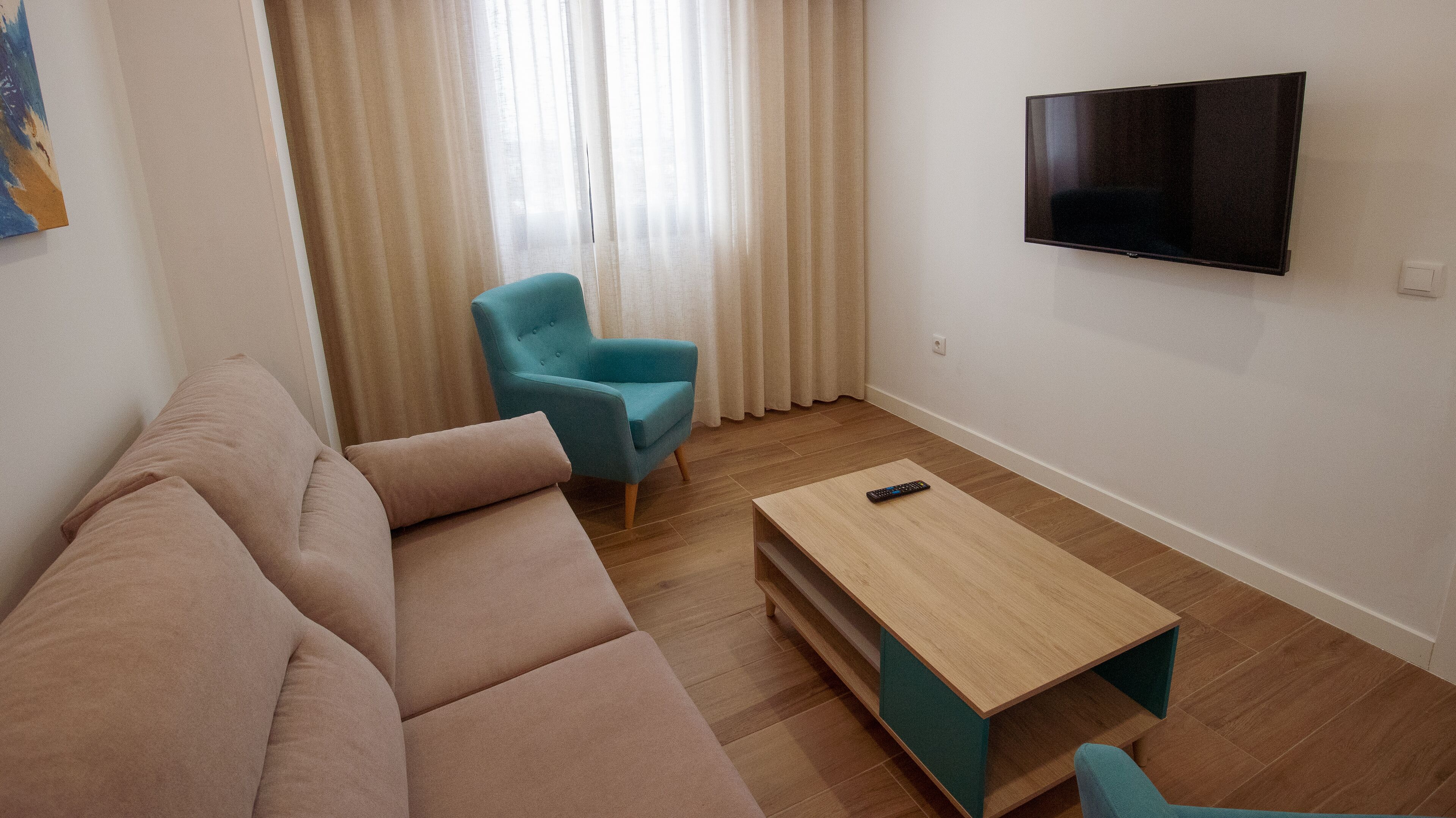 Een woonkamer met een bank, een teal fauteuil, een houten salontafel en een aan de muur bevestigde televisie.