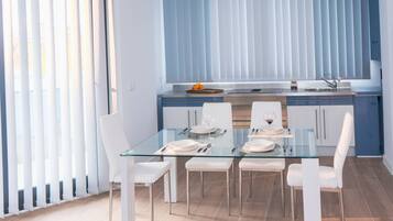 Una cocina moderna con una mesa de vidrio para cuatro personas, sillas blancas y una ventana con persianas.