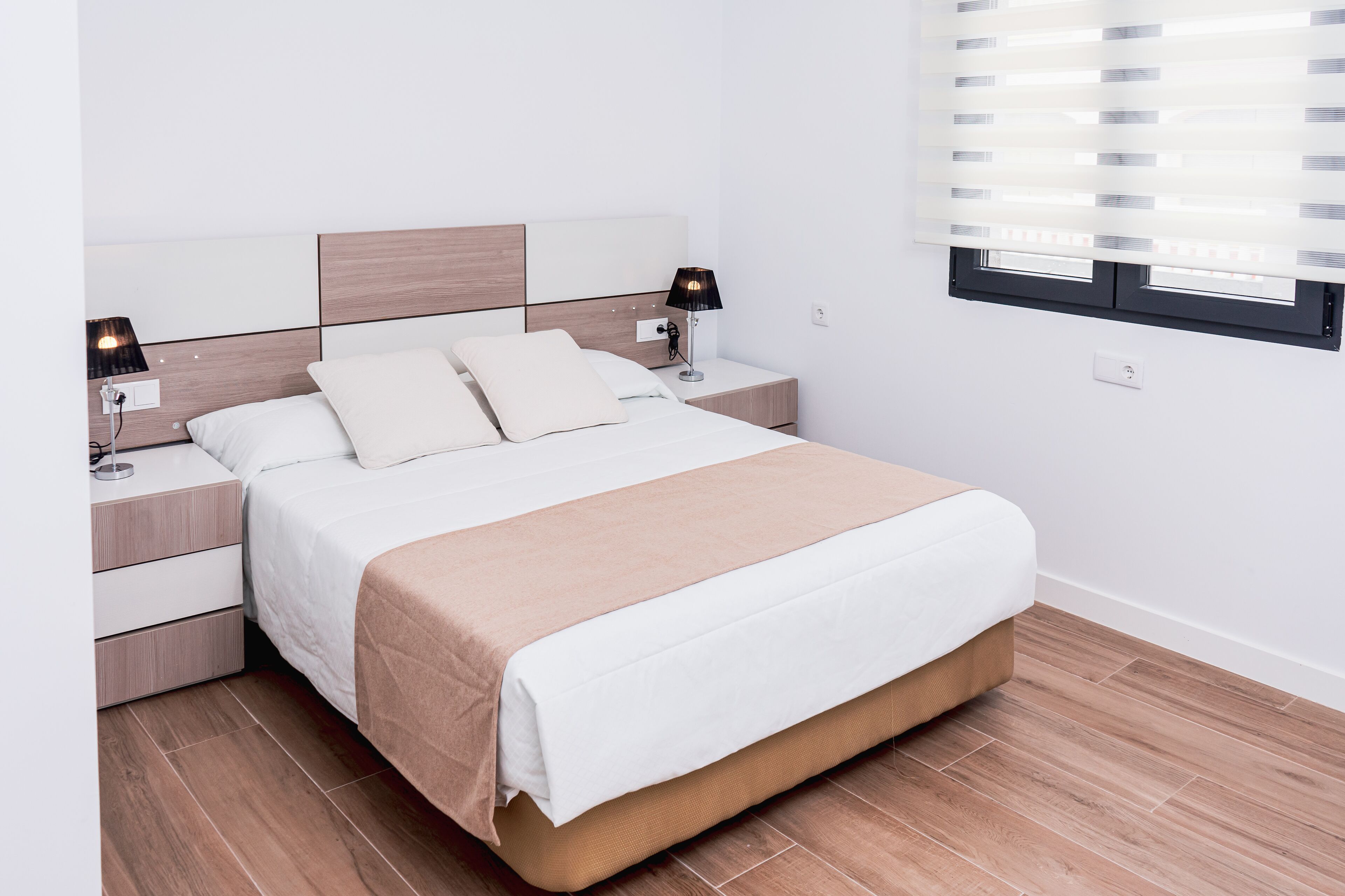 Un dormitorio moderno con una cama, dos mesitas de noche, un enchufe y una ventana con persianas.