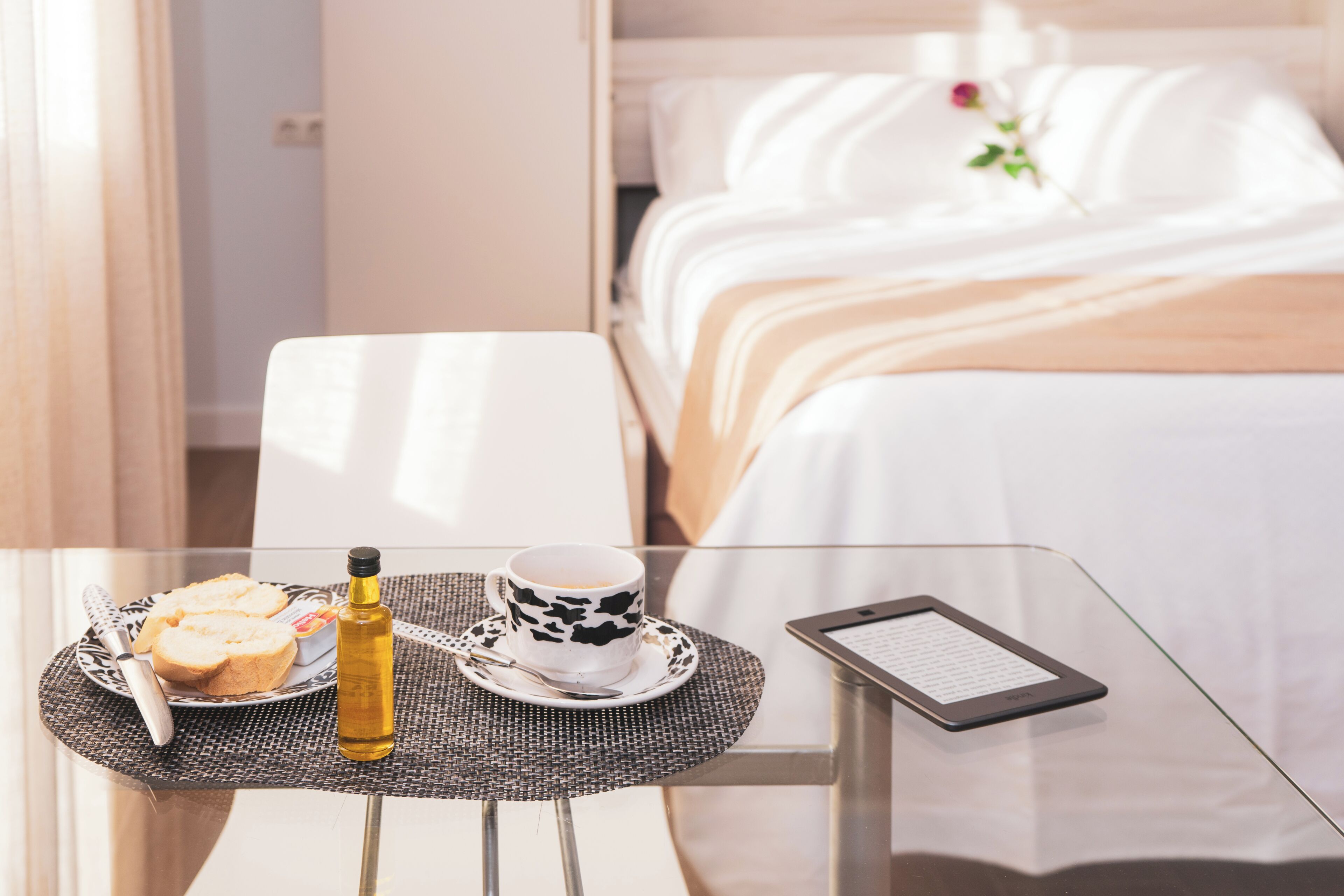 Habitación de hotel con una mesa de vidrio, una cama con ropa blanca, una mesita de noche con un libro y una flor sobre la cama.
