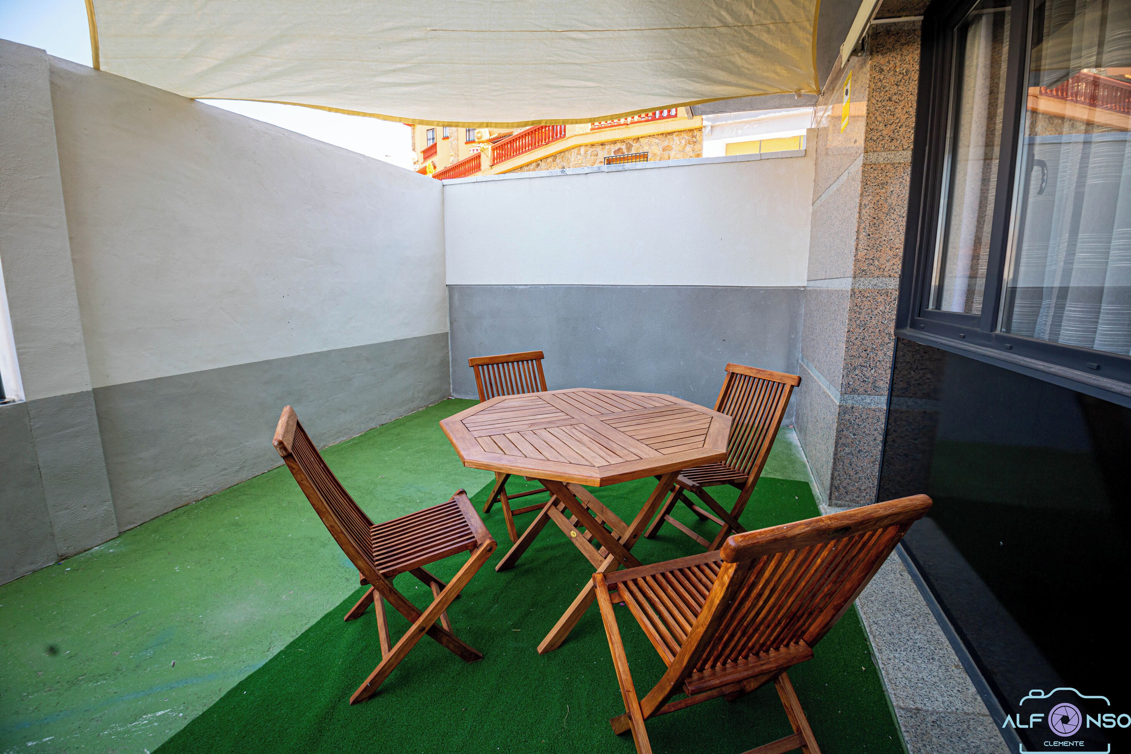 Un patio con una mesa redonda de madera y cuatro sillas, rodeado de un piso de césped artificial verde y paredes blancas.
