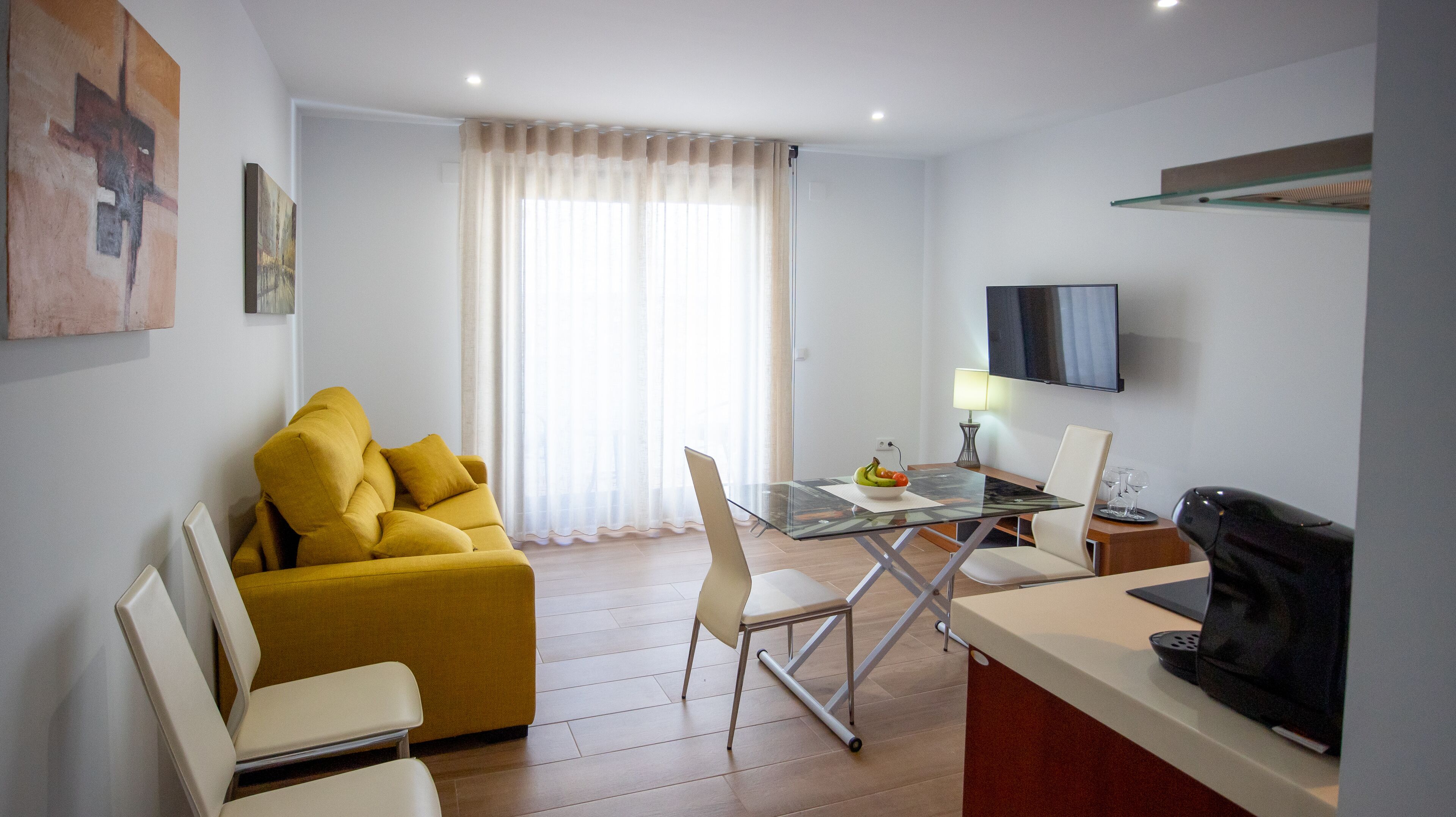 Una sala de estar moderna con un sofá amarillo, una mesa de comedor de vidrio y un televisor montado en la pared.