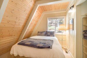 A cozy attic bedroom with a sloped ceiling, a bed with patterned bedding, a floor lamp, and a window with a view of the outdoors. - Pet Friendly with Big Views! (Tahoe City)