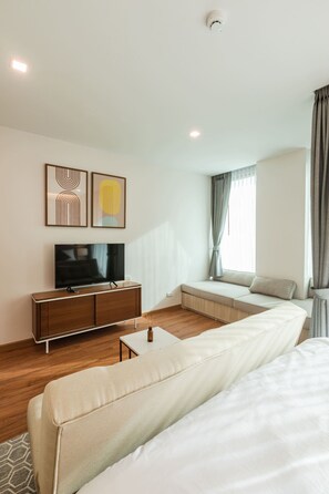 Una habitación de hotel moderna con una cama grande, un televisor sobre un mueble de madera, una zona de estar con sofá y cortinas, y obras de arte en la pared.