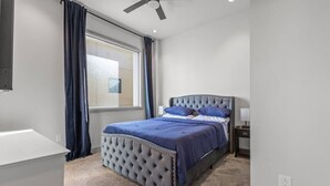 Un dormitori amb un llit amb capçaliner guarnicionat, una finestra amb cortines, un ventilador de sostre i una taula de nit amb una làmpada.