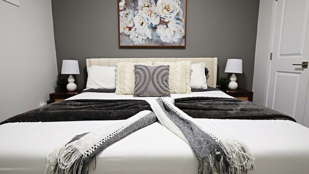 A neatly made bed with a white and gray blanket, two bedside lamps, and a floral painting on the wall. - Beautiful Suite King Bed|Long Stays|Disney|Airport (Edmonton)