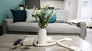 A living room with a grey sofa, a white vase with flowers, a remote control, and a white beaded string on a wooden coffee table. - Beautiful Suite King Bed|Long Stays|Disney|Airport (Edmonton)