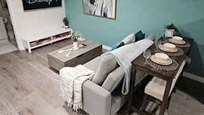 A living room with a grey sofa, a coffee table, a bookshelf, a dining table set with plates and glasses, and a Disney+ sign on the wall. - Beautiful Suite King Bed|Long Stays|Disney|Airport (Edmonton)