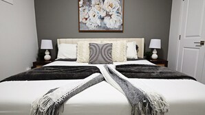Un lit bien fait, recouvert d’une couverture blanche et grise, avec deux lampes de chevet et un tableau floral accroché au mur.