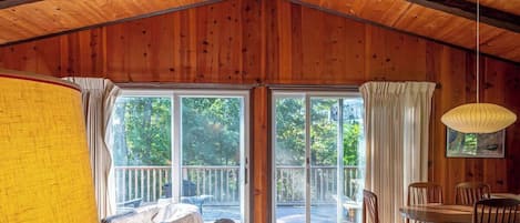 A cozy cabin interior with a wooden ceiling, a sofa, a dining table with chairs, and a large window with a view of the outdoors.