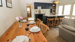 A dining area with a wooden table set for four, a vase with flowers, and a view into the kitchen with dark cabinets and a large island. - The Courtyard (Carlisle)