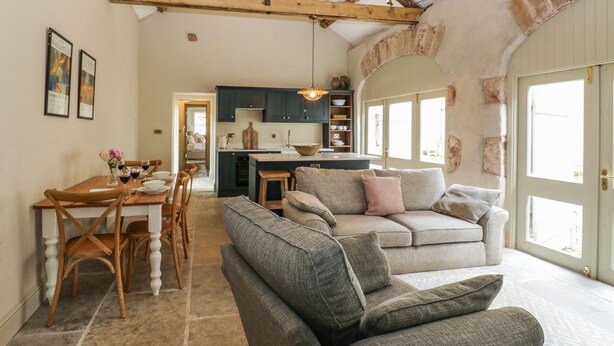 A cozy living room with a grey sofa, wooden dining table, and chairs. A kitchen with dark cabinets and a large window is visible in the background. - The Courtyard (Carlisle)