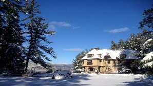 Ein schneebedecktes Haus mit Steinuntergrund und mehreren Fenstern, umgeben von schneebehafteten Bäumen und wolkenlosem, blauem Himmel.
