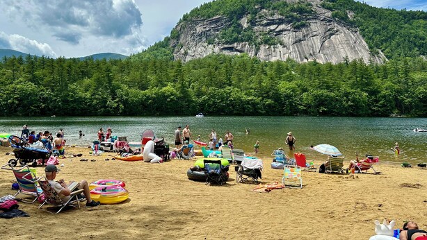 Ein Strand mit badenden und sonnenden Menschen, umgeben von üppiger Vegetation und einer hohen Klippe.