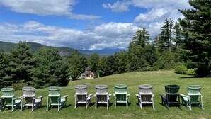 Eine Reihe Adirondack-Stühle auf einer grünen Wiese, im Hintergrund ein Haus und eine gebirgige Landschaft.