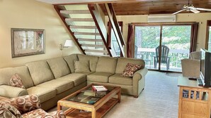 Ein Wohnzimmer mit einer Eckcouch, einem Glastisch, einem gemusterten Sessel, einem Holznachttisch mit einem Fernseher und einer Treppe, die auf einen Balkon mit Gartenmöbeln führt.