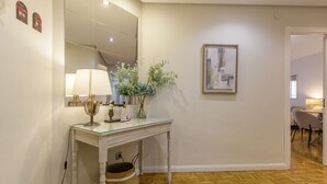 A room with a wooden floor, a white console table with a lamp, a vase with flowers, a framed painting on the wall, and a door leading to another room.
