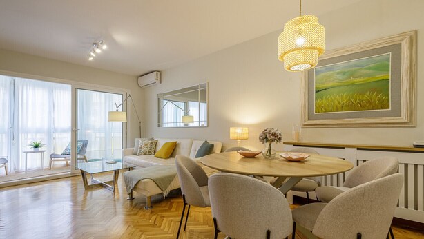 A modern living room with a round dining table, comfortable seating, a large window with sheer curtains, and a painting on the wall.