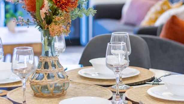 Ein Esstisch ist gedeckt mit weißen Tellern, klaren Gläsern und einer gewebten Tischmatte. Auf dem Tisch steht eine Vase mit orangefarbenen Blumen.