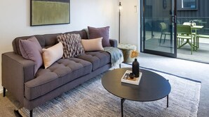 En moderne stue med en grå sofa, et rundt sofabord og en glasdør, der fører til et udendørs område med et bord og stole.