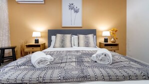 Ein ordentlich bezogenes Bett mit gemusterten Bettwäsche und zwei aufgerollten Handtüchern, flankiert von Nachttischen mit Lampen und Vasen, in einem Raum mit warmer Wandfarbe und einem gerahmten Bild über dem Bett.